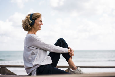 Smiling mature woman wearing wireless headphones sitting at beach