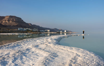 Distant view of woman standing on salt at dead sea against blue sky