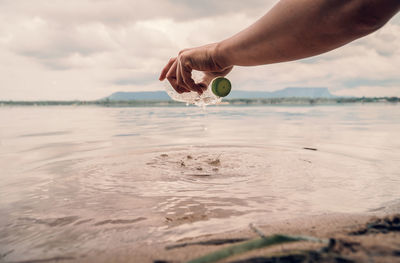 Cropped hand picking plastic bottle from sea at beach against cloudy sky