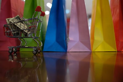 Close-up of colorful shopping bags with paper currencies in miniature shopping cart on wooden table