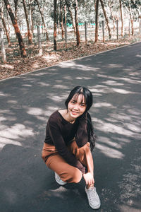 High angle portrait of smiling young woman crouching on road against trees in forest