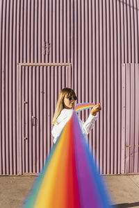 Young woman looking over shoulder holding rainbow colored ribbon in front of corrugated wall