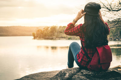 Rear view of woman sitting by lake against sky during sunset