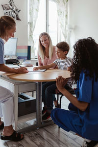 Doctor looking at patient pointing on paper while female nurse and mother sitting by desk in hospital