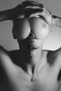 Close-up of shirtless man covering eyes with artificial boobs against wall at home