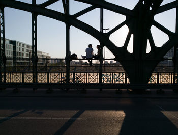 Silhouette people sitting on bridge in city against sky