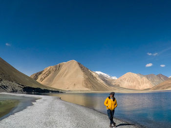 Full length of woman standing by lake against mountains and blue sky
