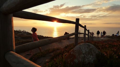 People on field by sea against sky during sunset