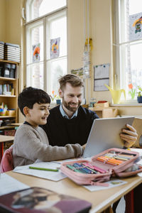 Happy male teacher assisting schoolboy using tablet pc at desk in school