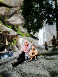 Low angle portrait of siblings sitting on rock against waterfall