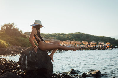 Side view of sensuous woman sitting on rock at beach during sunny day