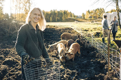 Portrait of smiling female farmer holding empty basket with pigs grazing at organic farm