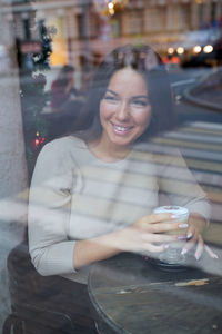 Smiling woman with coffee sitting in restaurant seen through window