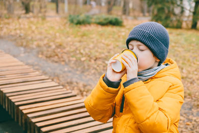 Boy in a yellow jacket drinks hot cocoa from a yellow glass, sitting on a bench, in the park.