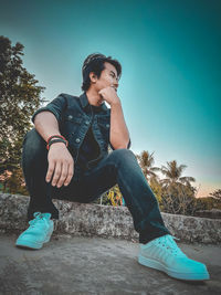 Young man looking away while sitting on tree against clear sky