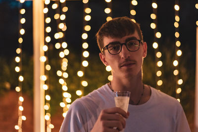 Portrait of young man holding drink while standing against illuminated lights at night