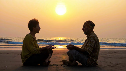 Men sitting on beach against sky during sunset