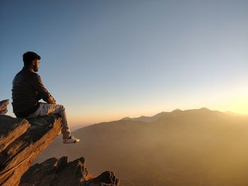 Man sitting on cliff against sky during sunset