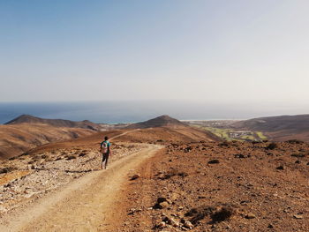 Rear view of man walking on dirt road against clear sky during sunny day