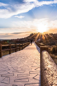 Footpath by railing against sky during sunset