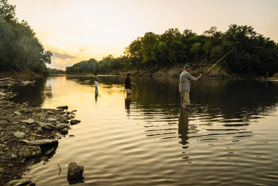 Friends enjoying fishing while standing in lake against sky during sunset