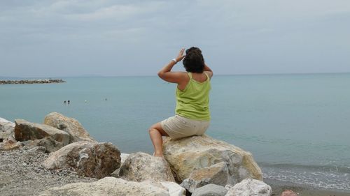 Rear view of woman sitting on rock at beach against sky