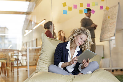 Woman in office using tablet in bean bag with meeting in background