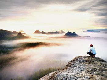 Hiker in white black clothes on cliff edge looking to misty hilly valley bellow, fall season begins