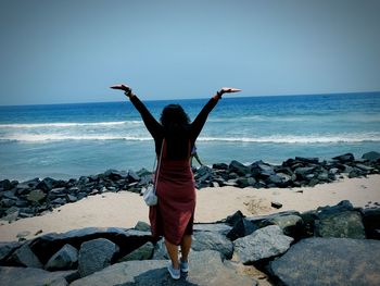 Rear view of woman with arms outstretched standing at beach against sky