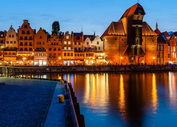 Illuminated buildings by river against sky at dusk