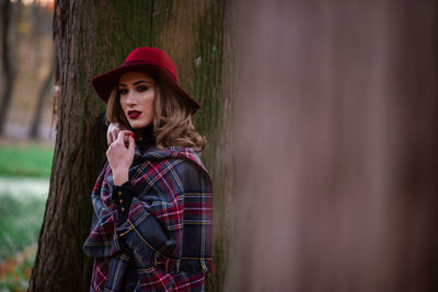 Portrait of woman wearing hat standing against tree trunk