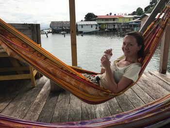 Portrait of smiling woman with drink resting in hammock by lake