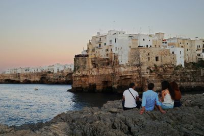 Rear view of friends sitting on rocks at sea shore by buildings against clear sky during sunset