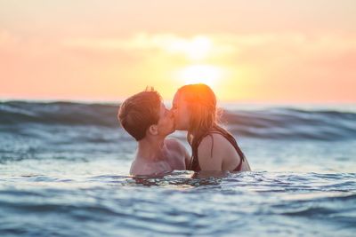 Young couple kissing in sea against sky during sunset
