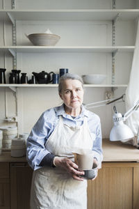 Portrait of senior female potter standing with pitcher against shelves at workshop