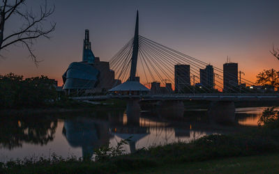 Bridge over river against sky during sunset with cityscape silhouette