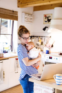 Mother with baby in kitchen looking at laptop