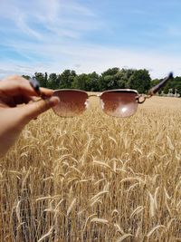 Cropped hand holding sunglasses on field against sky