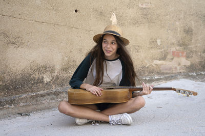 Young woman wearing hat playing guitar sitting on street