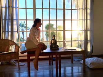 Woman sitting by buddha statute on table against window at home