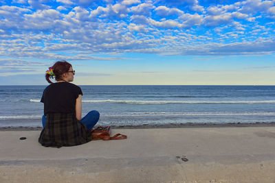 Rear view of woman sitting at beach against sky during sunset