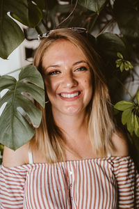 Young happy blonde woman smiling through the leaves of a monstera