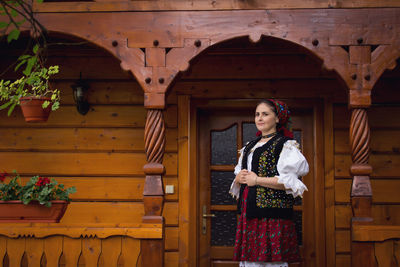 Thoughtful young woman in romanian clothing standing on porch of wooden house