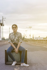 Portrait of young woman in sunglasses sitting on television against sky