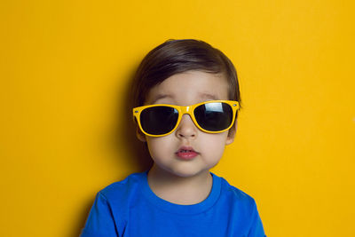 Cheerful baby boy in blue t-shirt stands on yellow background in sunglasses