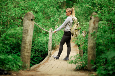 Side view of young woman with backpack standing on boardwalk in forest