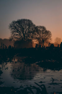 Silhouette bare trees by lake against sky during sunset