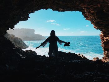 Woman standing on rock by sea against sky holding sandals