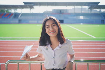 Portrait of smiling young woman holding paper airplane while standing in stadium