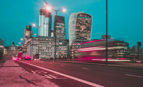 Illuminated city street by buildings against sky at night
